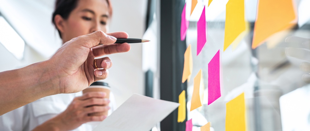 Hand mit Stift zeigt auf Glaswand mit bunten Klebezetteln