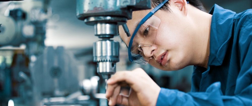 Nahaufnahme eines jungen männlichen Praktikanten mit Schutzbrille an einem Industriebohrer.