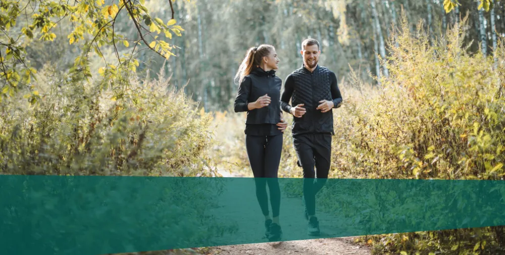 Ein junger Mann und eine junge Frau joggen in einem herbstlichen Wald und unterhalten sich freudig.